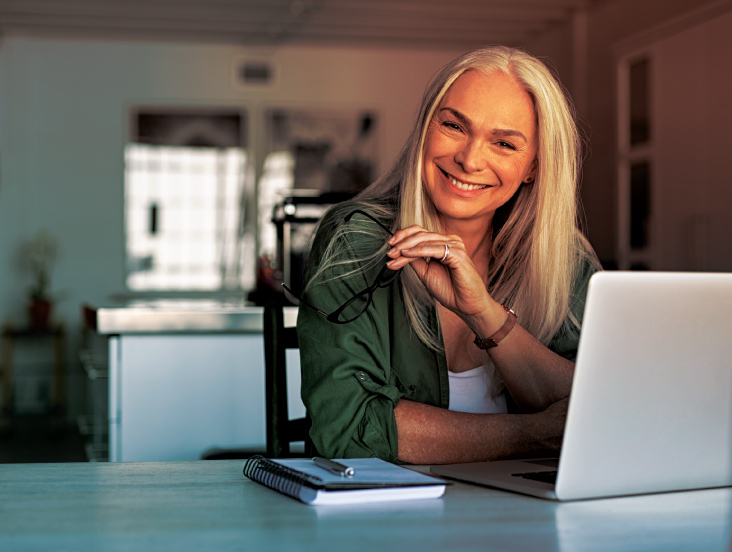 A smiling woman with long gray hair sits at a desk holding glasses in one hand. She is in a well-lit room with a notebook, pen, and laptop in front of her. She wears a green shirt over a white top.